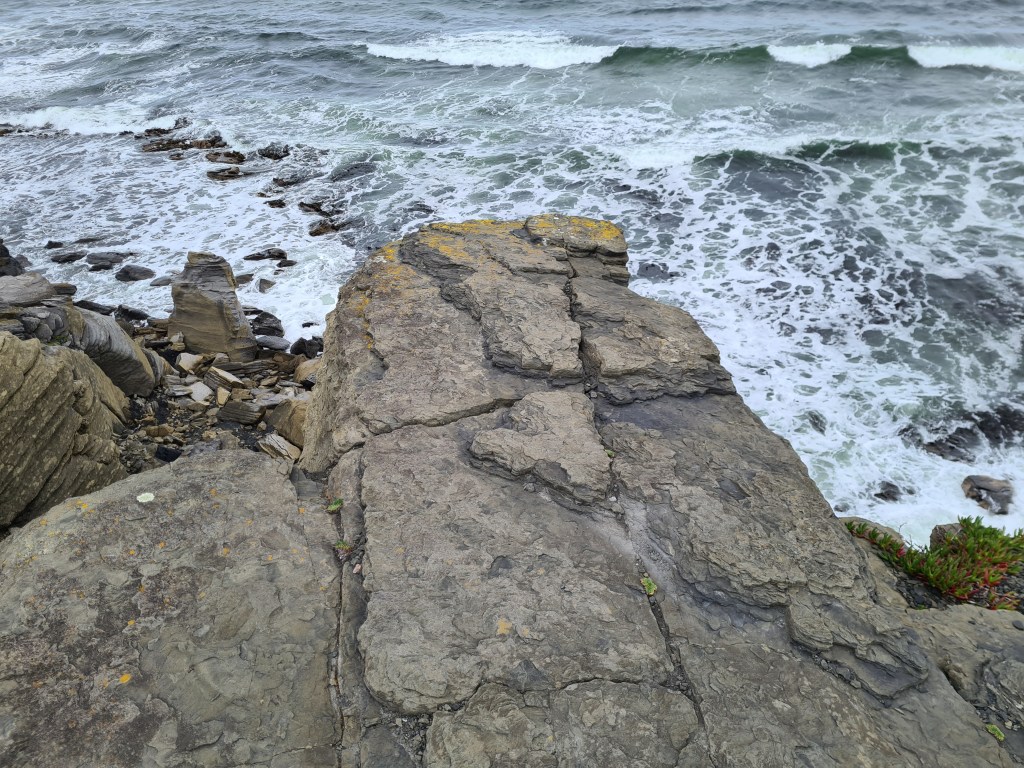 the foam of the waves make white veins through the dark blue and turquoise of the ocean. A jutting, cracked rock is in the way as the photographer was unwilling to walk to the true edge of the cliff.  