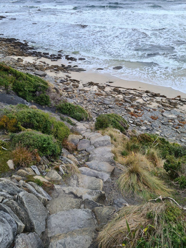 A stairway made of rocks leads down to a rocky beach. The tide is coming in and there is very little soft sand to see. 