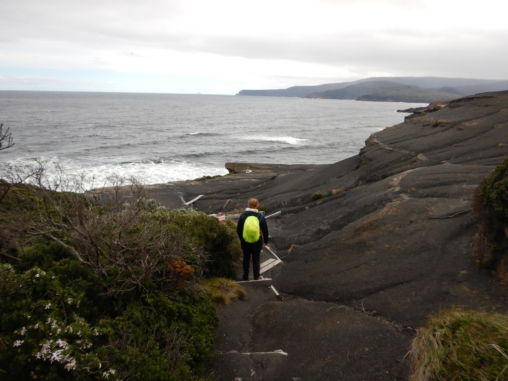 Sapphire, dressed in dark colours with a brightly coloured waterproof cover over her backpack admires the black cliffs of South Cape Bay. 