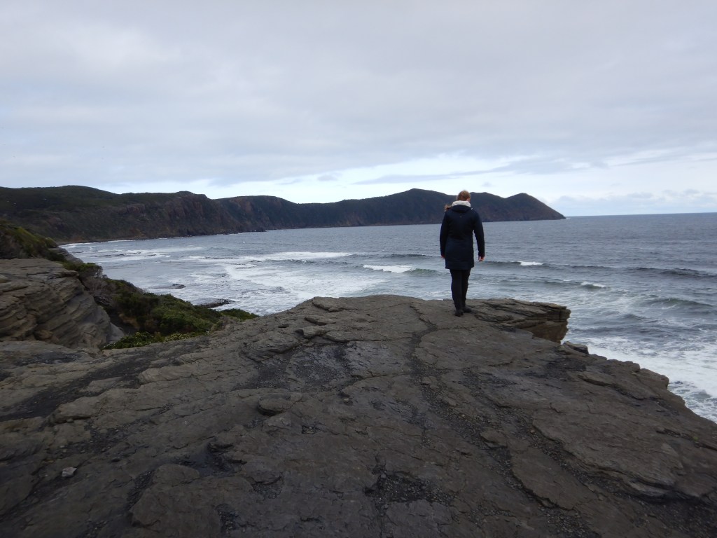 Sapphire is dressed in dark colours that match the black of the cliffs and deep blue of the ocean and distant peaks of the bay. She is standing near the cliff's edge. 