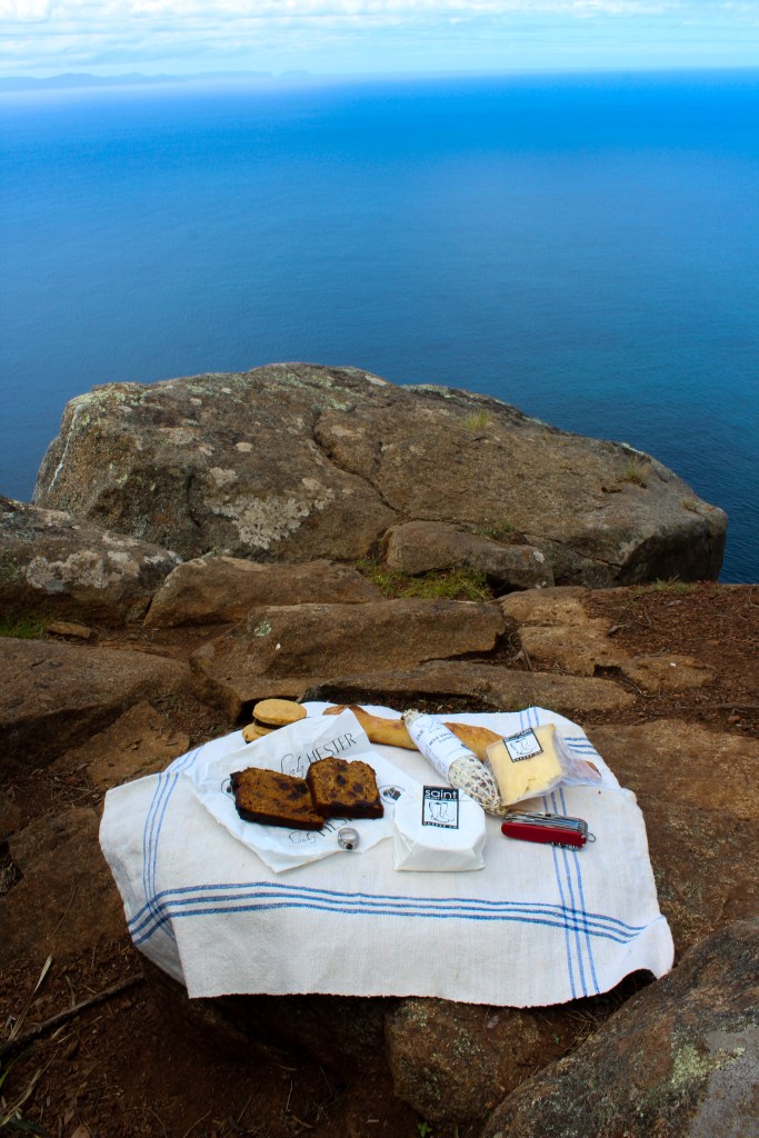 The picnic gathered earlier in the article is spread on a linen tea towel across a rock at the edge of the cliff.