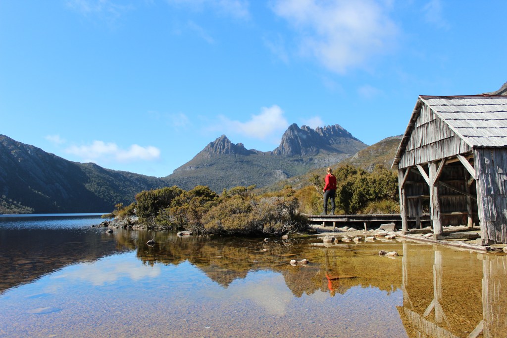 Sapphire is wearing a red shirt and walking along the raised path behind the dove lake boat house looking towards Cradle Mountain
