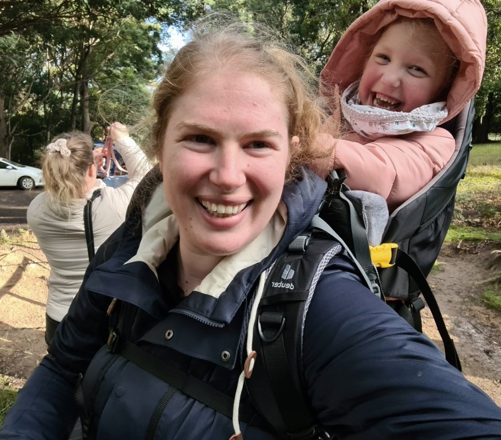 Sapphire, dressed in a navy blue raincoat carries her 4 year old client, who is wearing pink, on her back in a We Carry Kevan backpack. Both are smiling