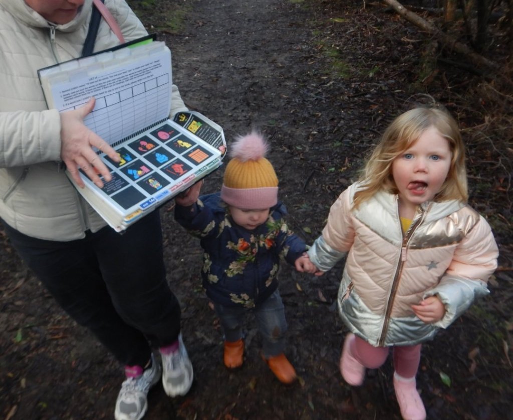 Mummy L holds the PODD book that we use to communicate as a family open while scanning to find the right word. The two youngest children hold on to her, very nervous about heading into the woods.