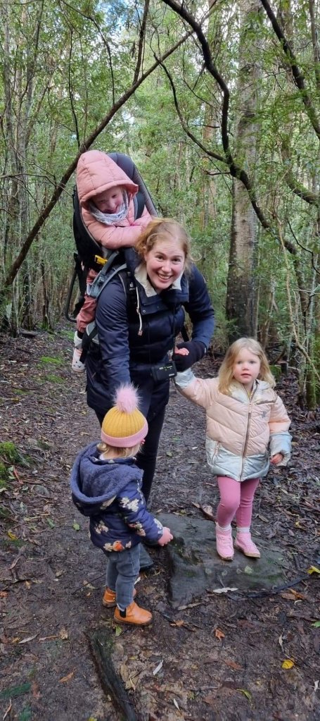 Sapphire, carrying Miss 4yo on her back in a We Carry Kevan pack, and hold the hands of the youngest 2 children. The picture is blurry due to the movement of the children, but Sapphire is smiling.