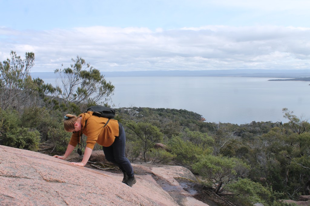 Sapphire is on all fours attempting to gain purchase of the rock. The ocean spreads out under the cliff below her.