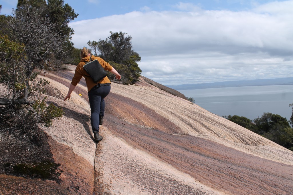 Sapphire holds her arms out from balance while following the very narrow crease across a particularly slippery, section with a steep drop into the ocean visible in the top right corner.
