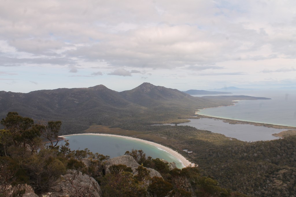 2 other mountain peaks are visible on the other side of an isthmus. One side of the isthmus is a white sand bay, the other is bushland.