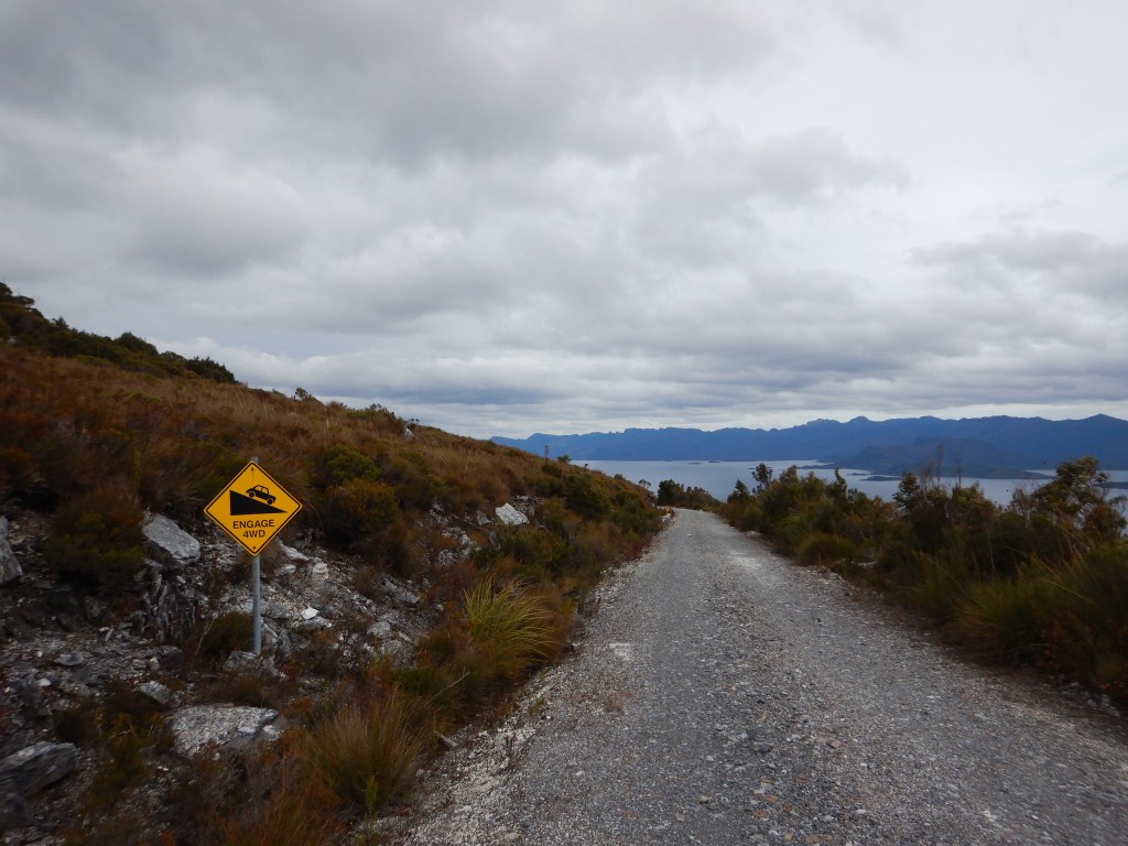 A grey gravel road dissappers over the edge of a hill and around a corner. Lakes and mountains are visible in the distance. A yellow warning sign reading "engage 4WD" is in the foreground. 