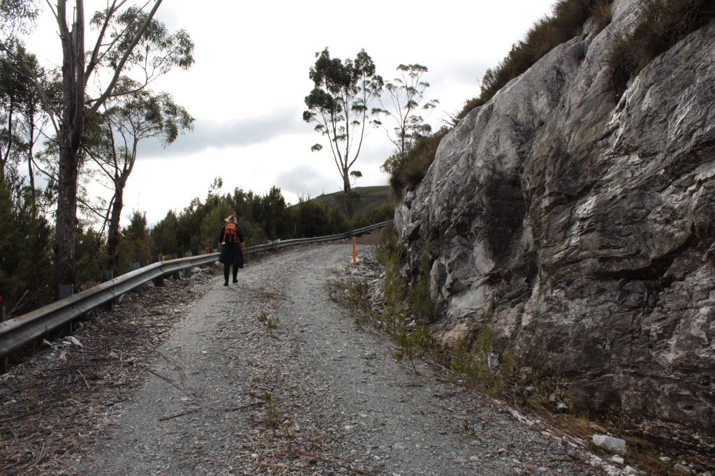 Sapphire, dressed in dark clothing and carrying a bright orange backpack walks along the broad gravel road. A steep rocky outcrop stands to the right of her, trees and a cliff are to the left. the road rounds a corner ahead of her. 