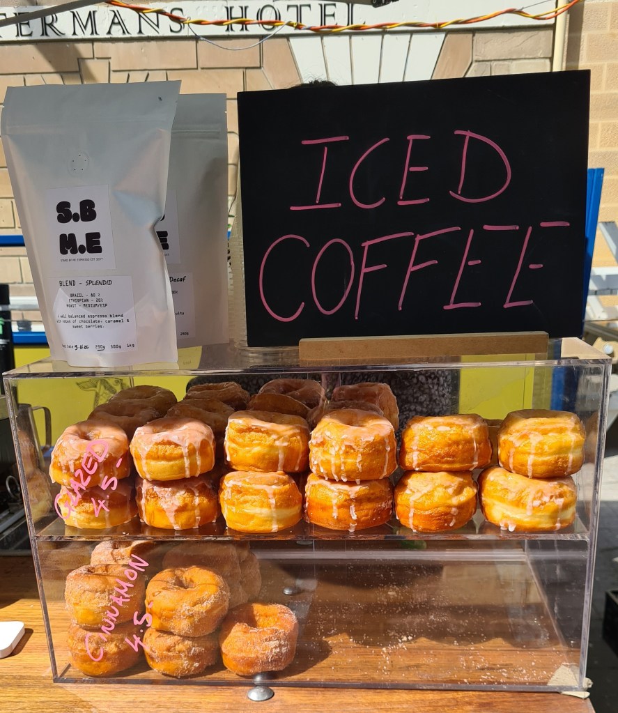 Glazed and sugared donuts sit in stacks in a display cabinet, a large sign the reads "Iced coffee" in pink writing on a blackboard sits above them. 