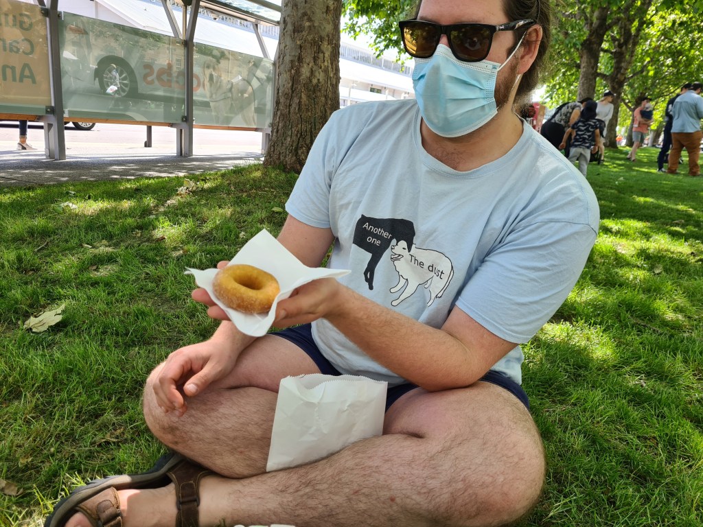 Sommelier sits crossed legged on the grass holding a donut on a serviette. 